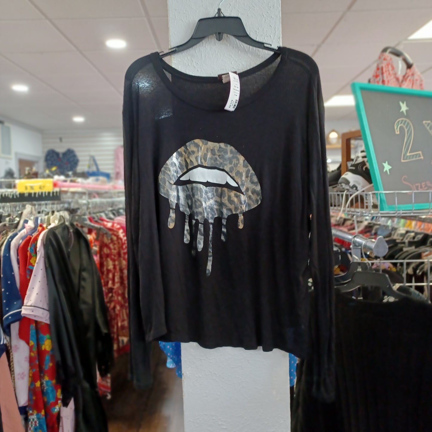 A black long-sleeve top hanging on a hanger in a clothing store, featuring a leopard-print dripping lips graphic on the front.